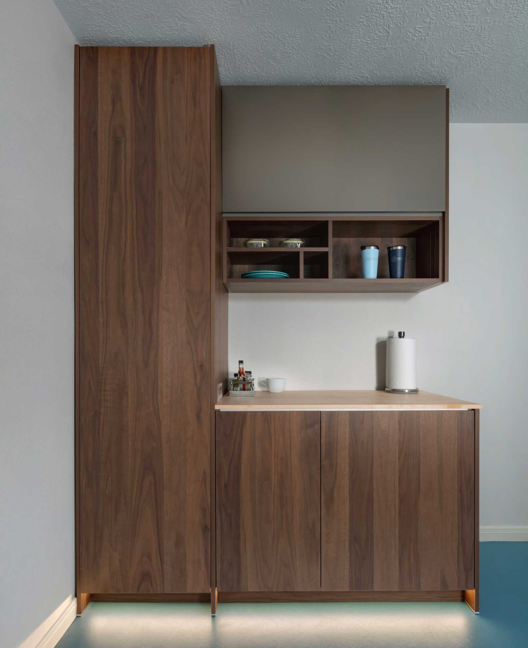 Minimalist kitchen corner with dark wood cabinets, open shelves holding bowls and cups, a few condiments, a paper towel roll, and under-cabinet lighting.