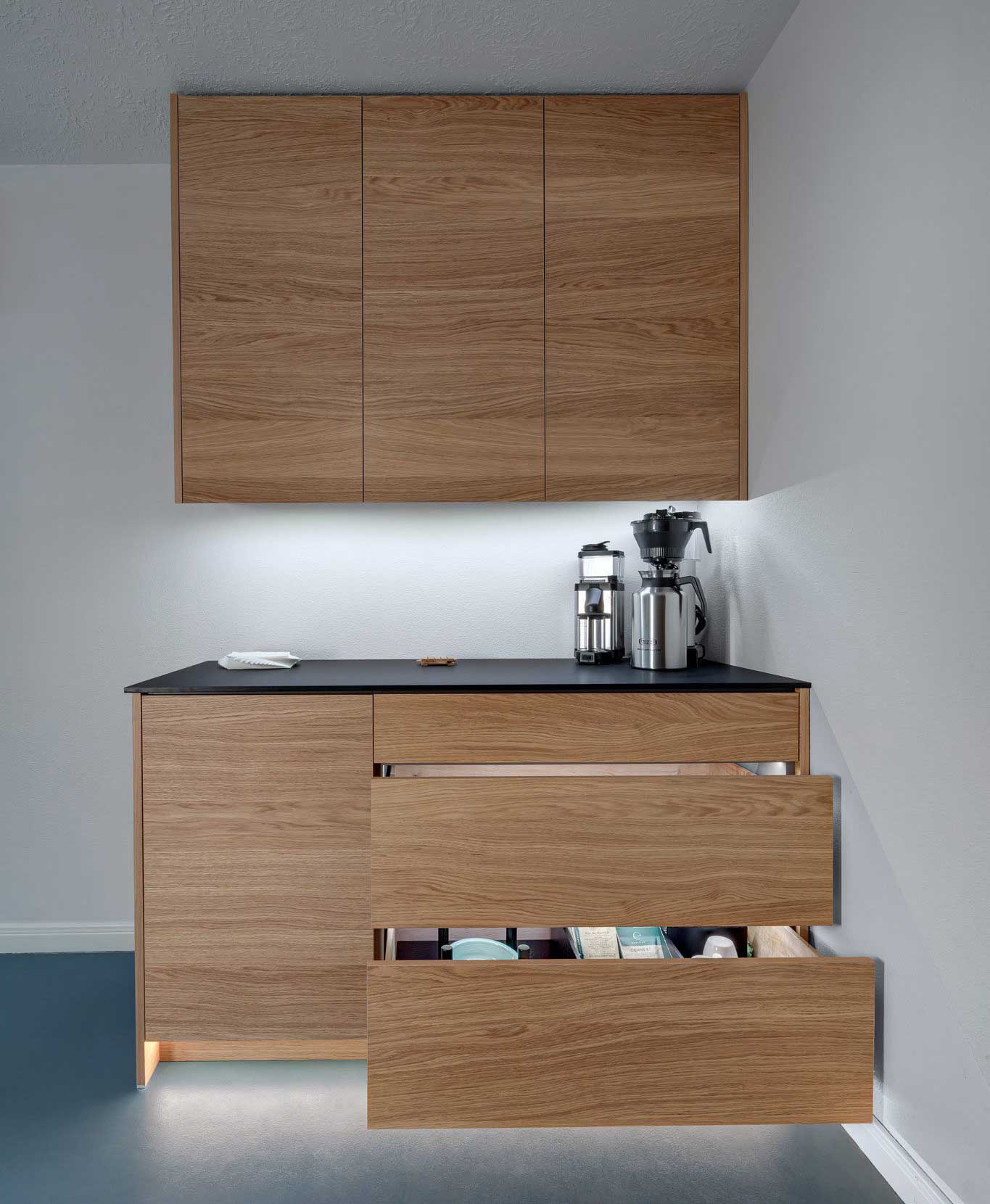 Modern kitchen with wooden cabinets, black countertop, and two open lower drawers. Coffee maker and mixer sit on the counter under wall-mounted cabinets.