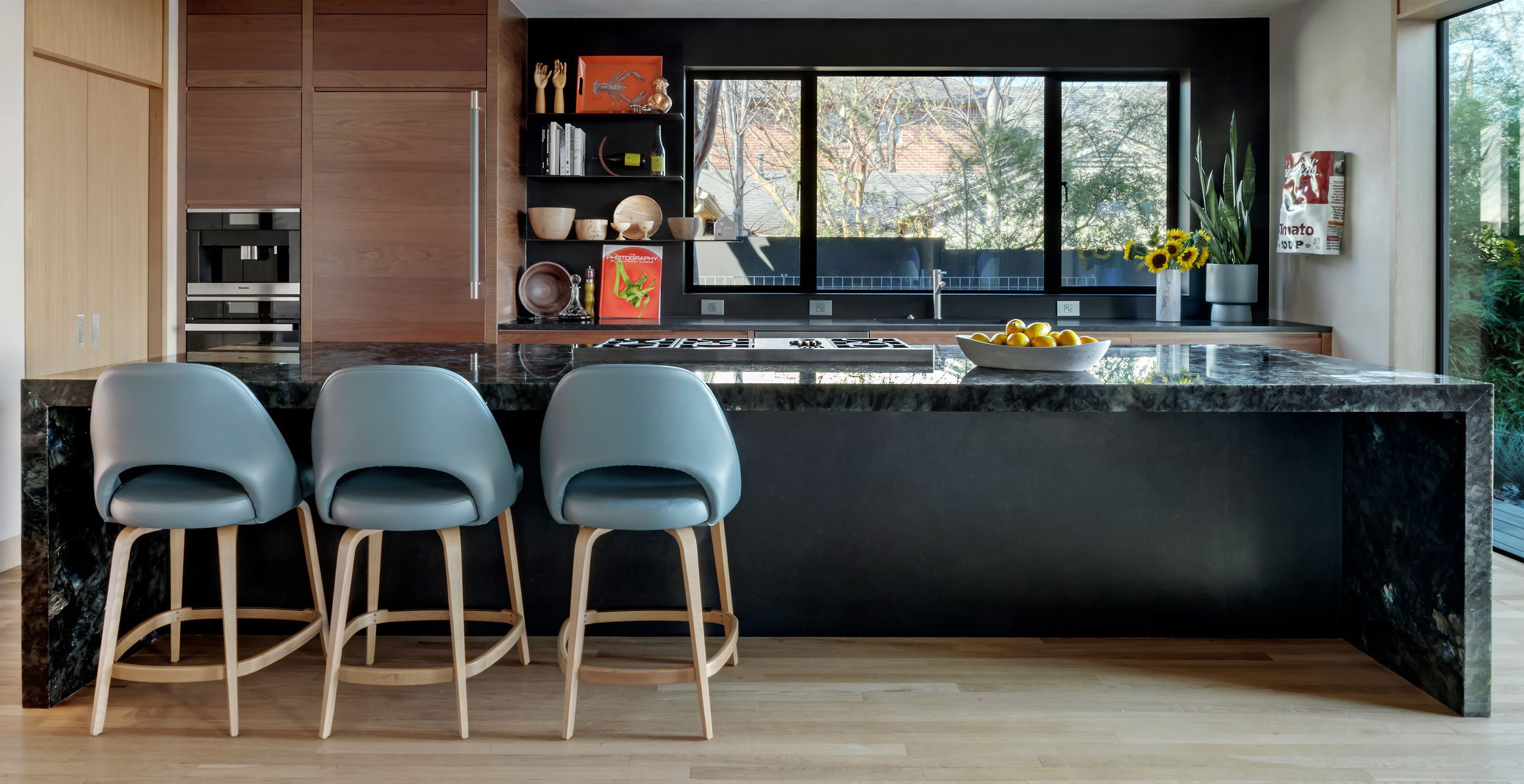Modern kitchen with a dark marble island, three blue barstools, wooden cabinets, open shelves, a bowl of lemons, and large windows overlooking trees.